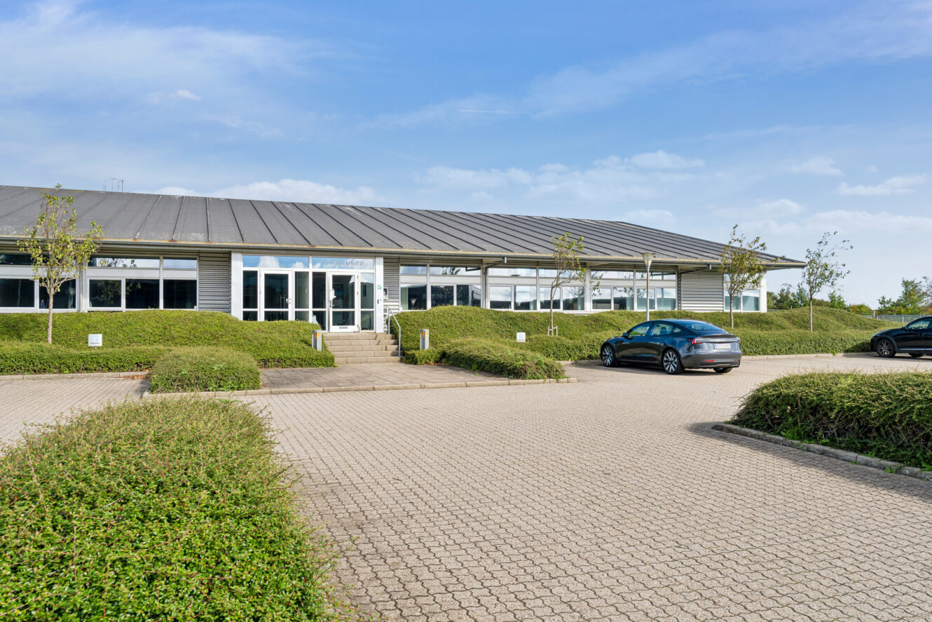 En moderne kontorbygning i én etage med store vinduer, et metaltag og en stort set tom parkeringsplads med to parkerede biler og klippede buske.