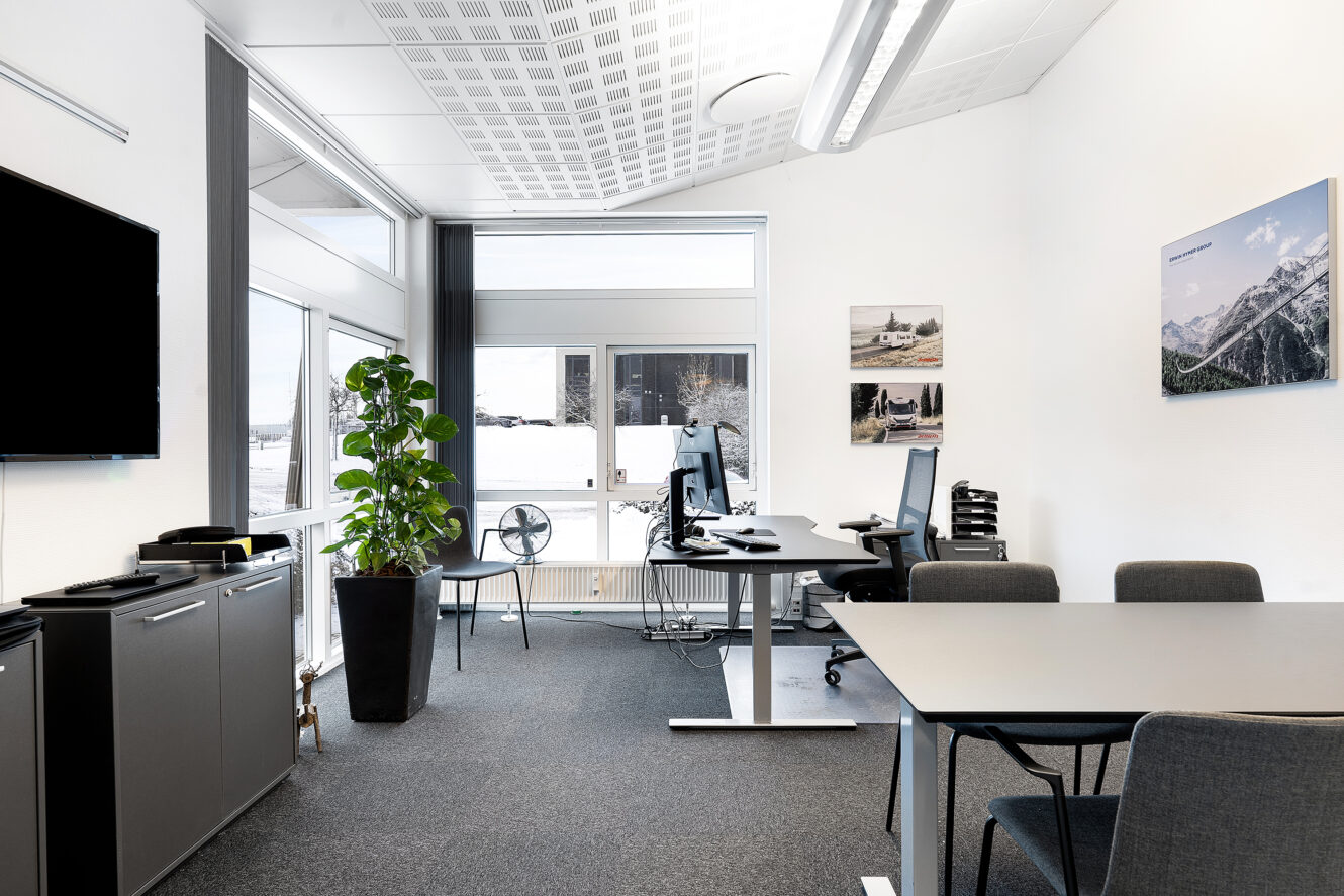 Moderne kontor med store vinduer, et skrivebord med computerskærme, et bord med stole, en potteplante, vægkunst og et tv på venstre væg.