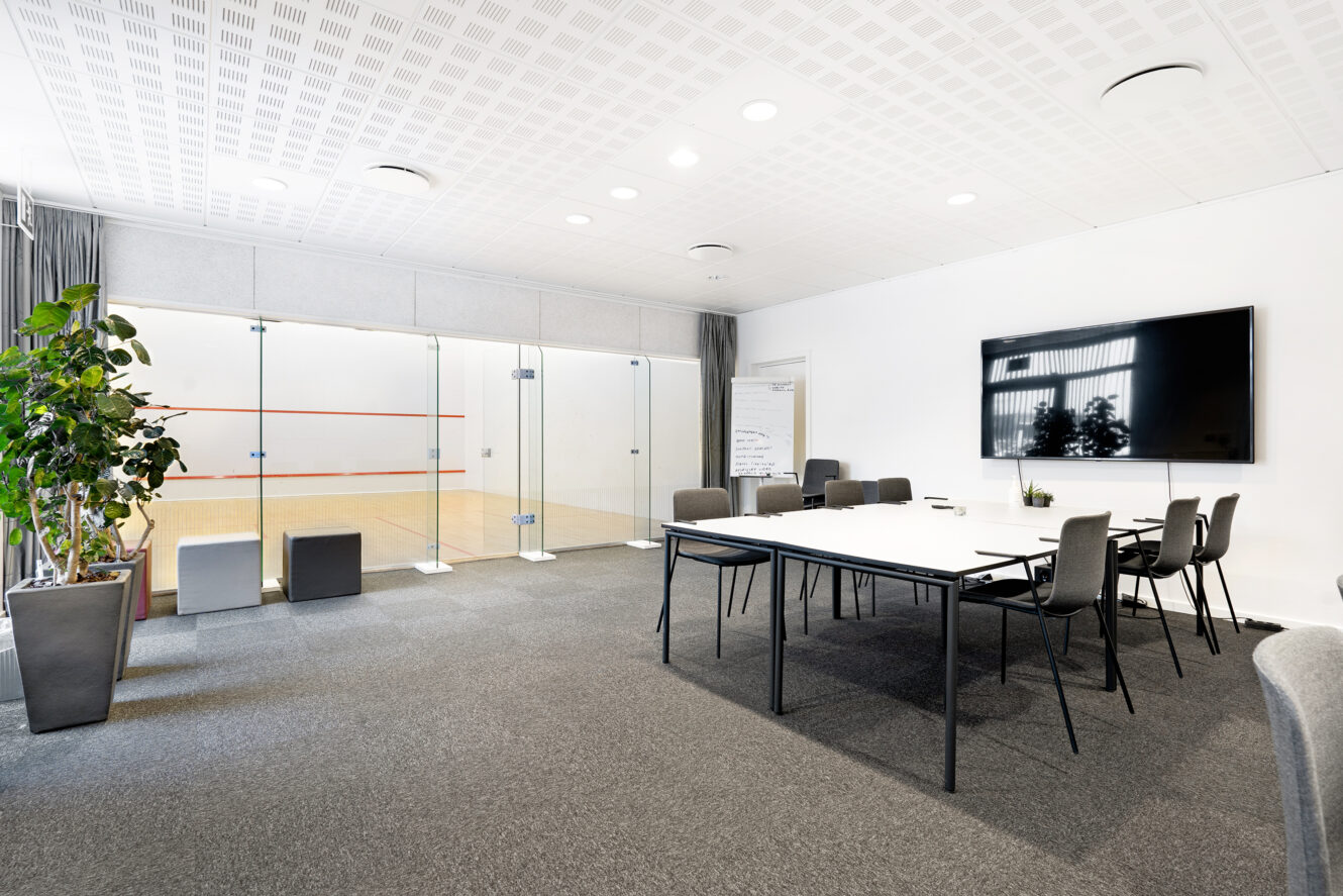 Et moderne konferencelokale med bord, stole, en stor skærm og en glasvæg, der adskiller det fra en squashbane. En potteplante og grå siddekuber er også til stede.