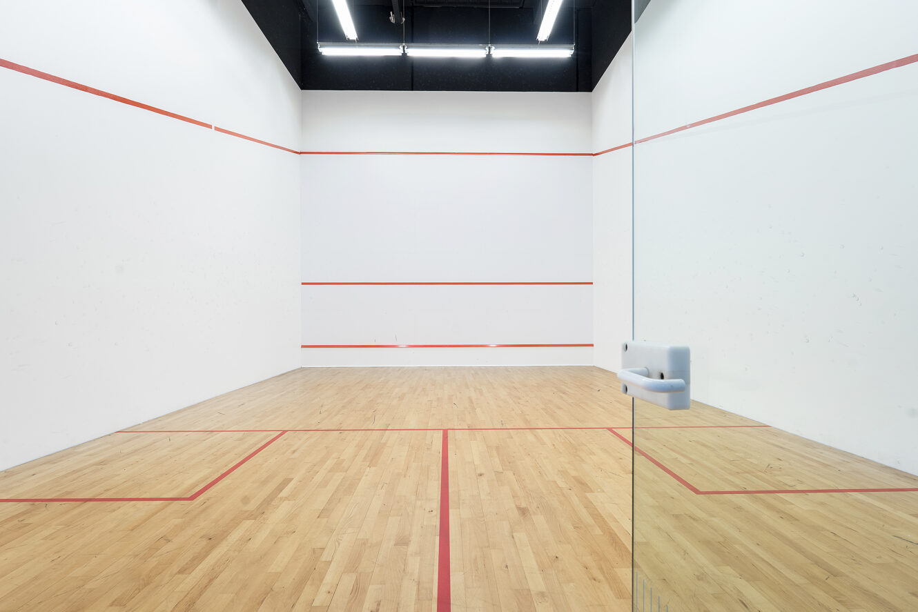 En squashbane med trægulv, hvide vægge markeret med røde grænselinjer og en delvist åben glasdør i forgrunden.