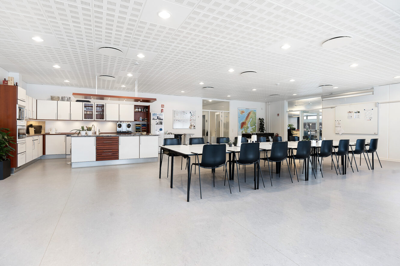 Rummeligt, moderne køkken og spiseplads med to lange borde, sorte stole, hvide skabe og god belysning.