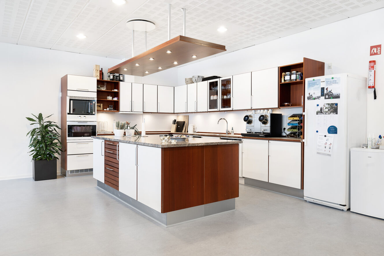 Moderne køkken med hvide skabe, trædetaljer, en central ø, indbyggede apparater, et køleskab og forskellige køkkenting på bordplader og hylder. En potteplante står i hjørnet.