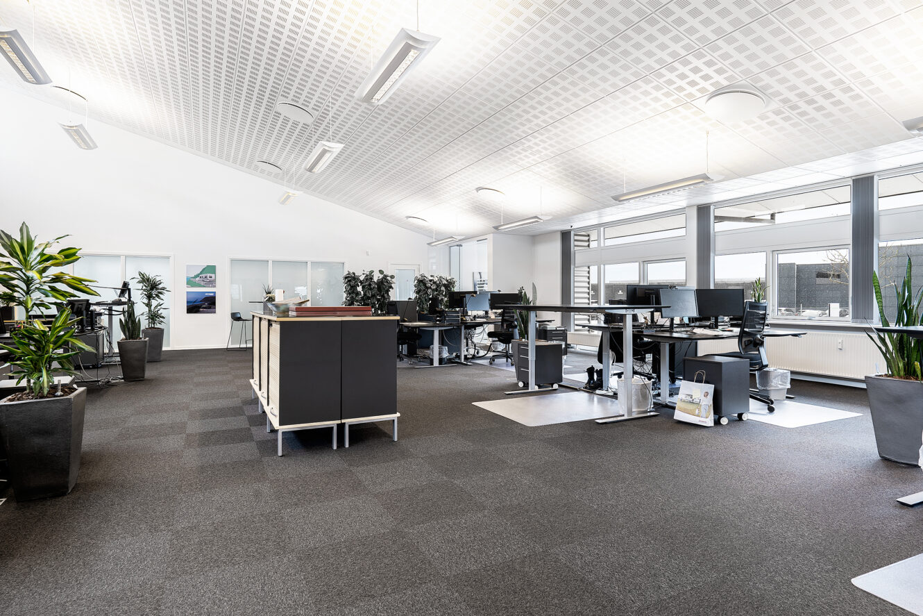 Moderne åbent kontorlandskab med store vinduer, flere skriveborde, computerskærme, ergonomiske stole og potteplanter, med god belysning og et neutralt farveskema.