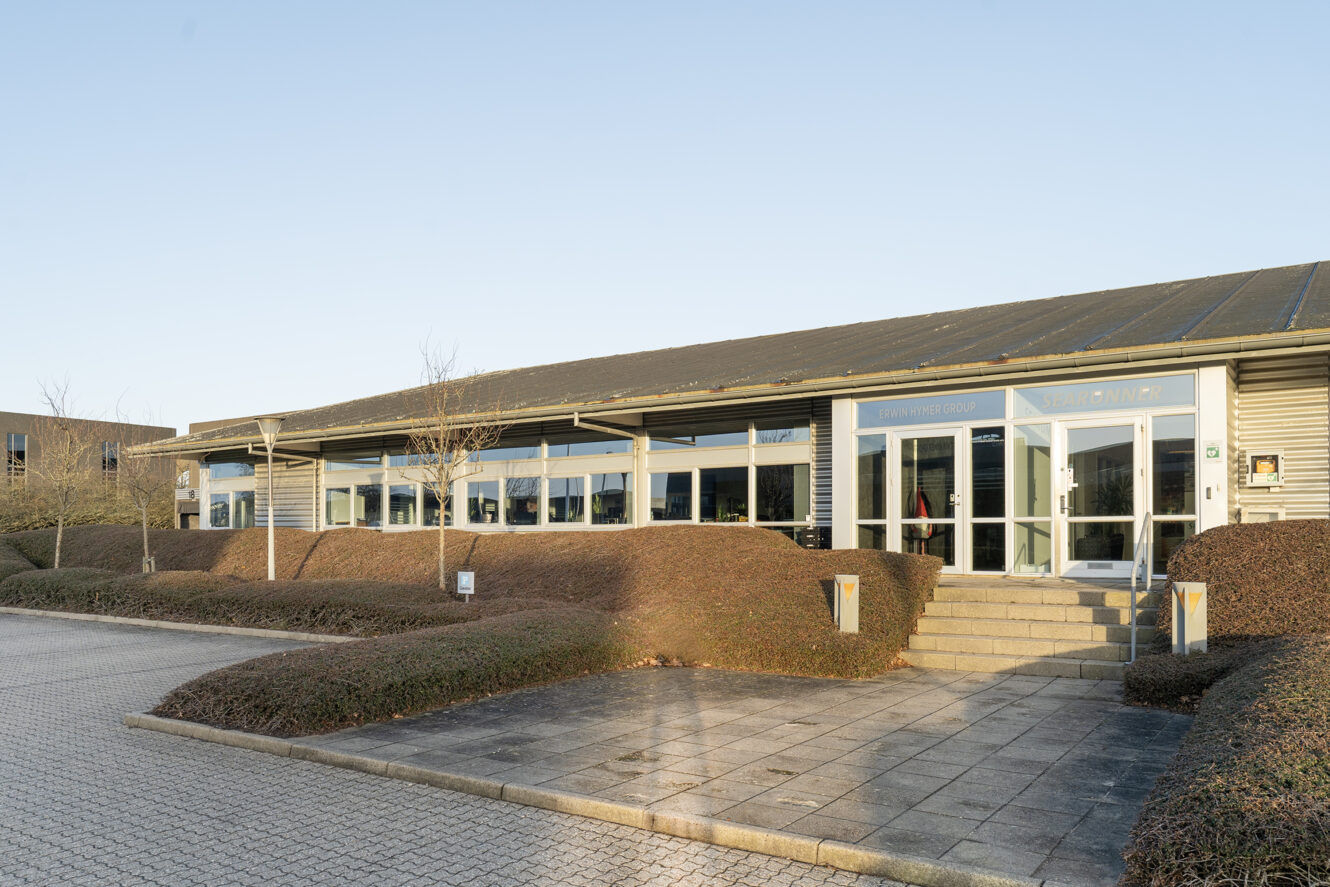 En erhvervsbygning i én etage med store vinduer, fladt tag og beskåret buskads foran, beliggende ved siden af en asfalteret gangsti og et parkeringsområde.