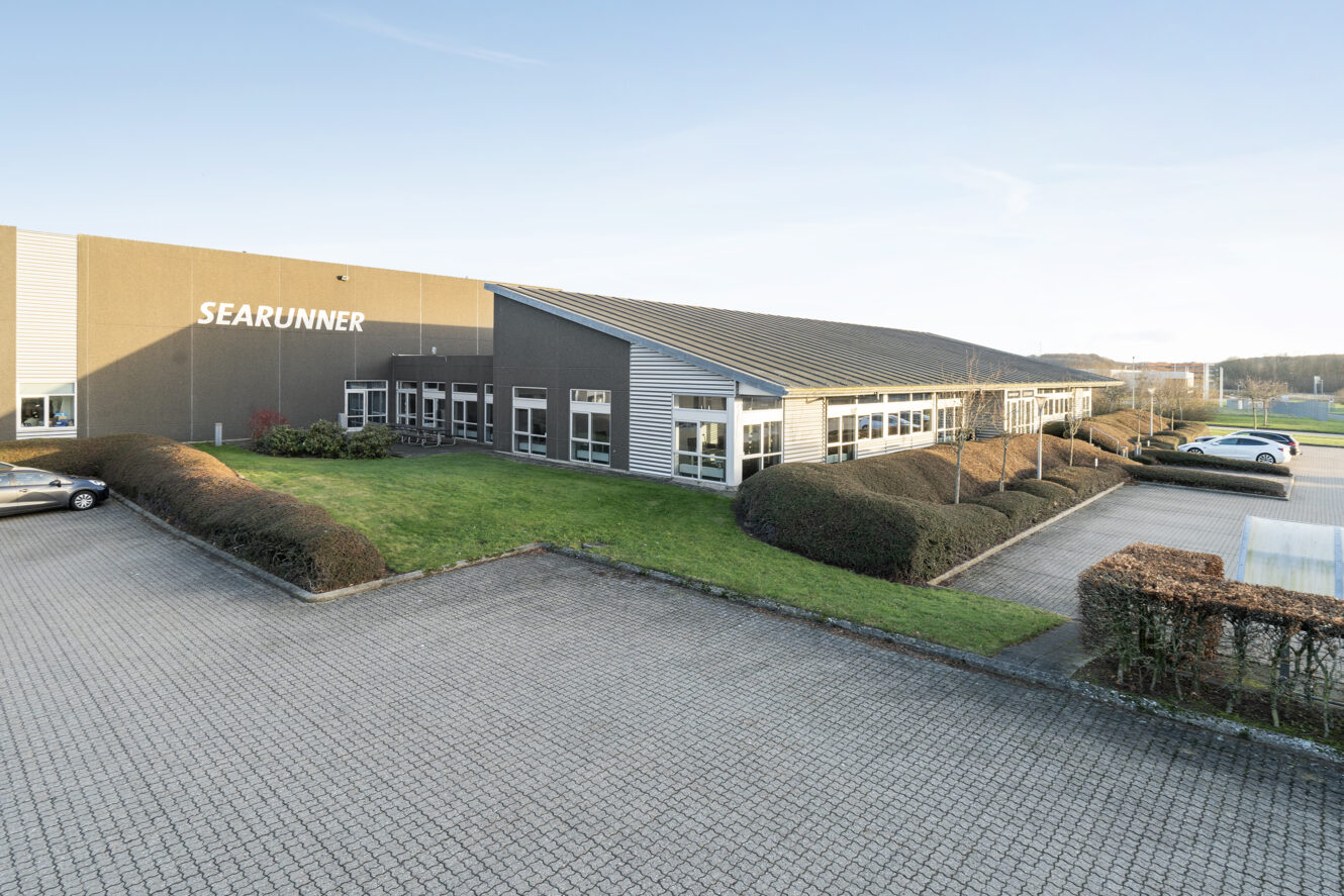 En moderne industribygning med navnet SEARUNNER og en pænt anlagt indgang, omgivet af parkerede biler og asfalterede områder under en klar himmel.