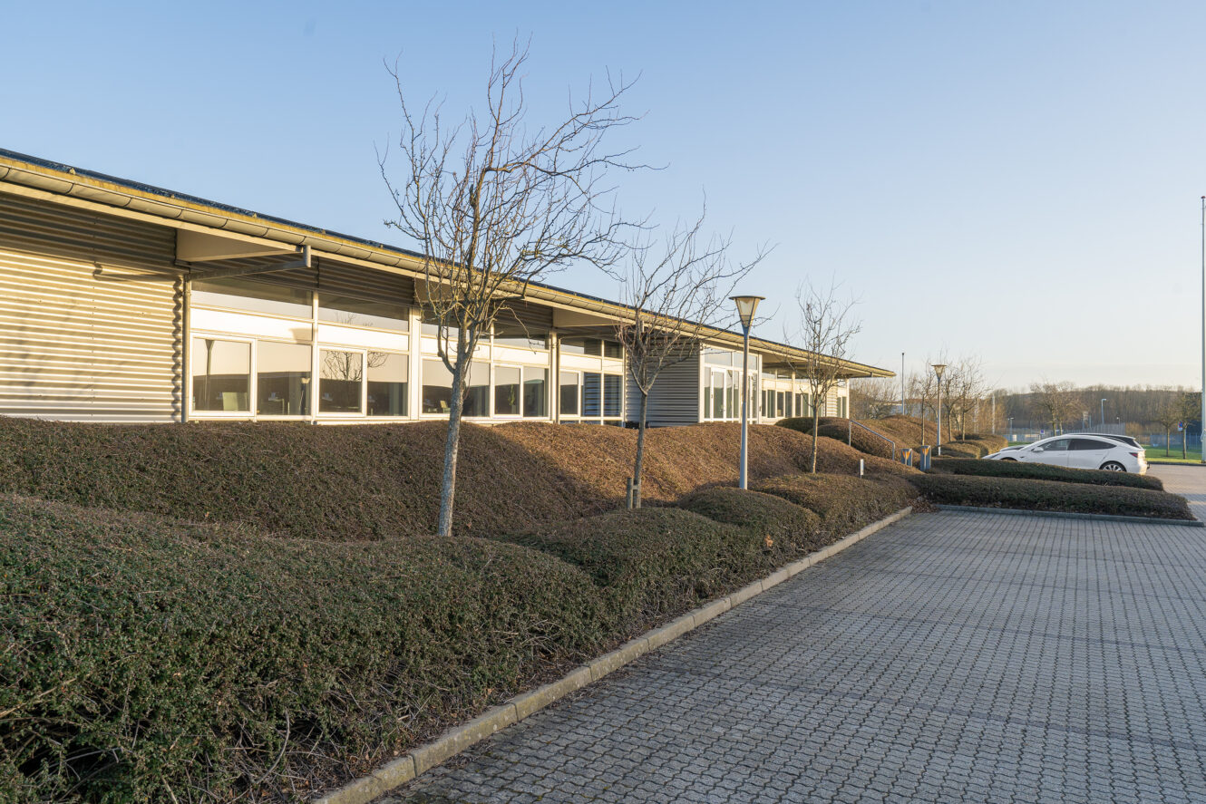 En moderne lav bygning med store vinduer, sirligt trimmede buske, bladløse træer og en tom asfalteret parkeringsplads under klar himmel.
