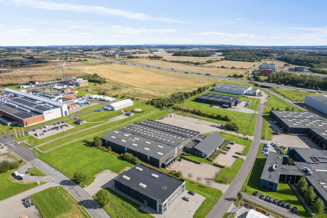 Luftfoto af en industripark med flere store bygninger, lagerbygninger, parkeringspladser og omkringliggende grønne marker nær en motorvej.