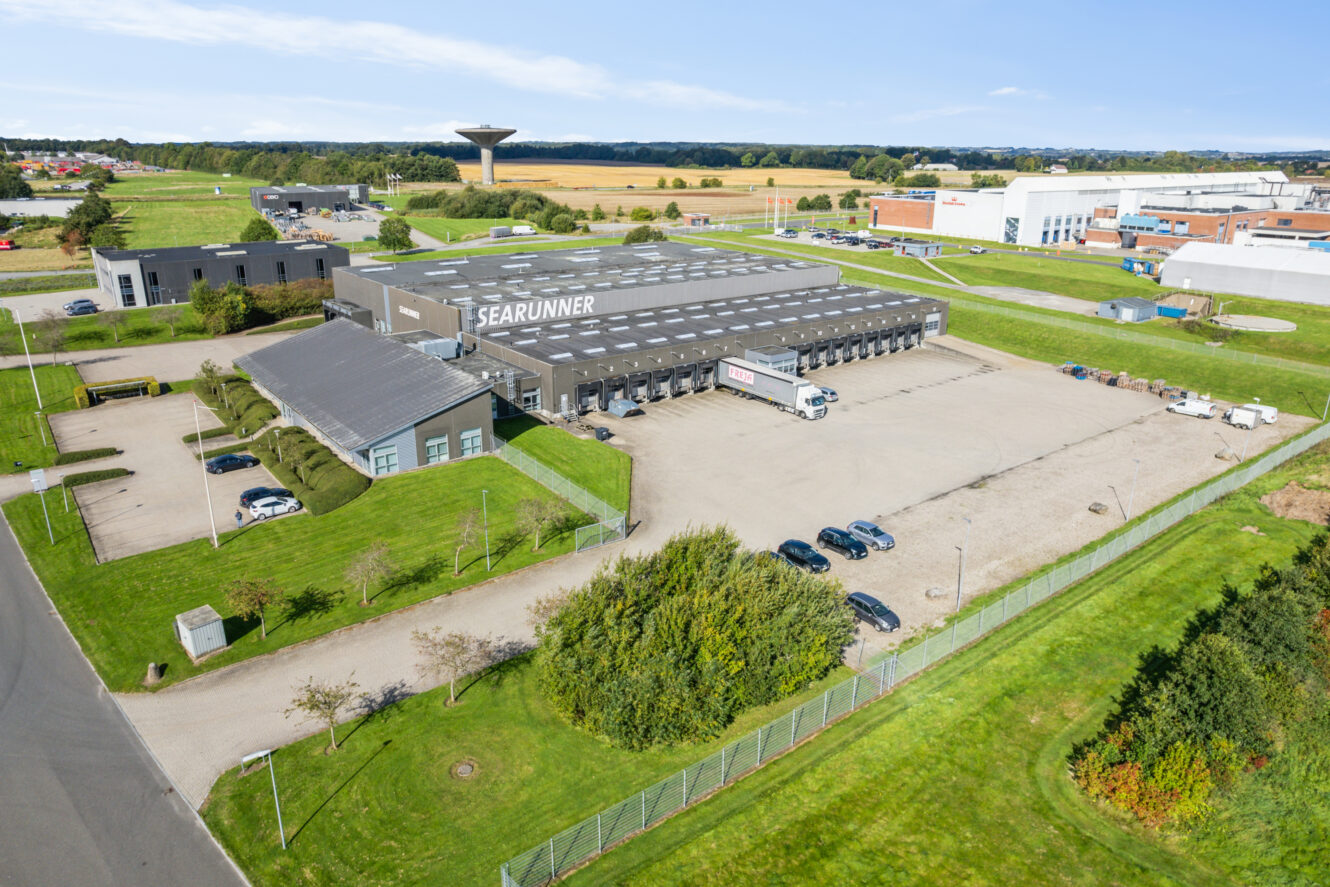 Luftfoto af et stort industrilager mærket SEARUNNER med en parkeringsplads, varevogne og omkringliggende grønne områder.