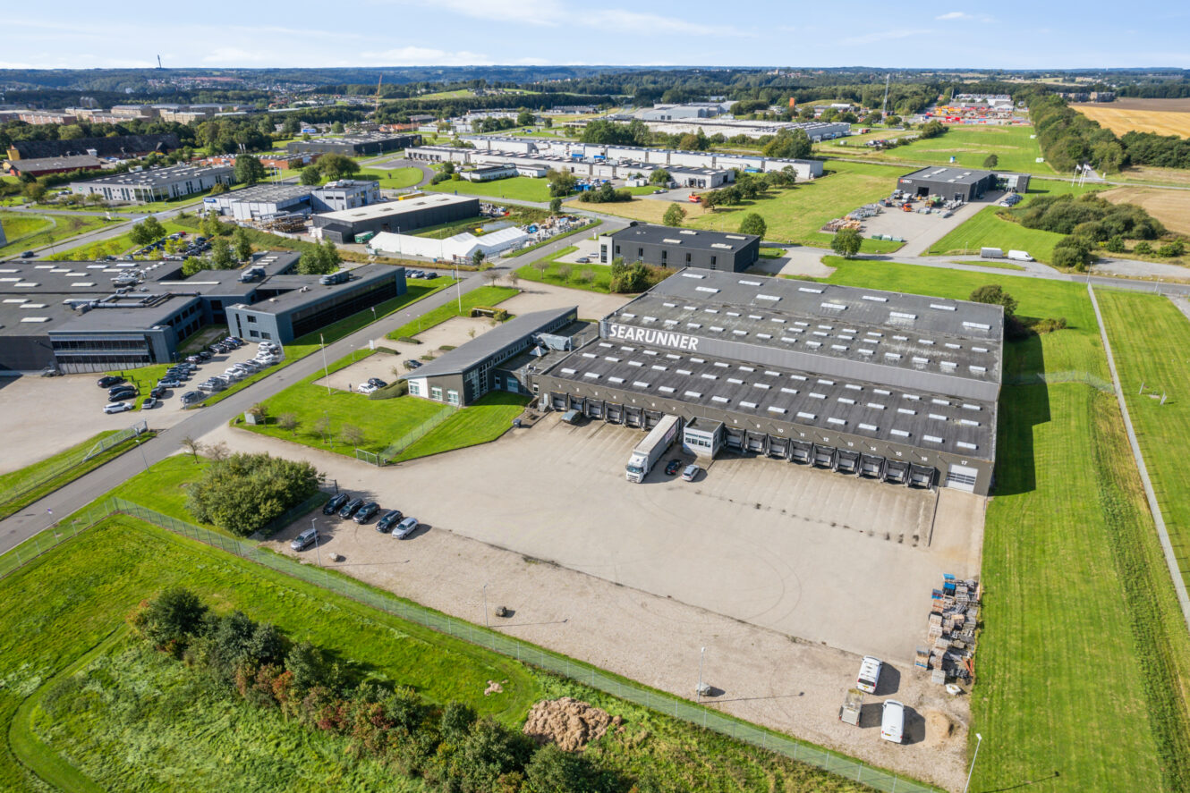 Luftfoto af et stort industrilager mærket SEARUNNER med lastbiler og biler parkeret udenfor, omgivet af grønne marker og andre industribygninger.
