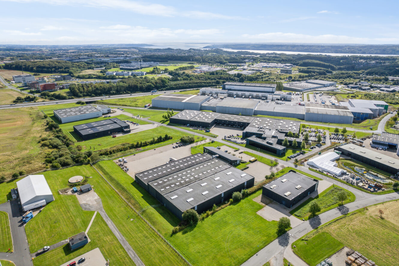 Luftfoto af en industripark med flere store lagerbygninger, grønne plæner, veje og omkringliggende marker under en delvis overskyet himmel.