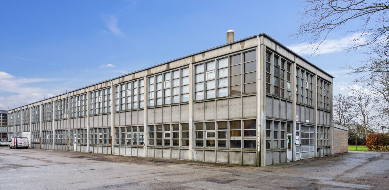 En grå industribygning i to etager med store gittervinduer, der ligger på en asfalteret grund under en blå himmel.