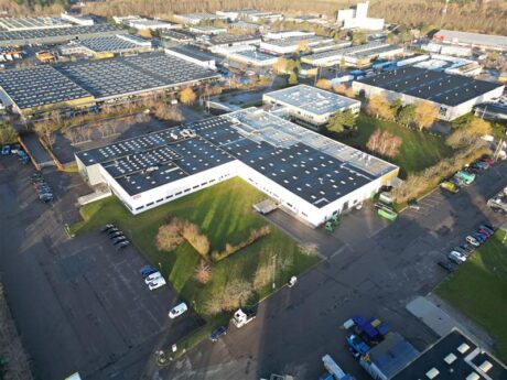 Luftfoto af et industrikompleks med flere store lagerbygninger, parkeringspladser, køretøjer og grønne områder på en solrig dag.