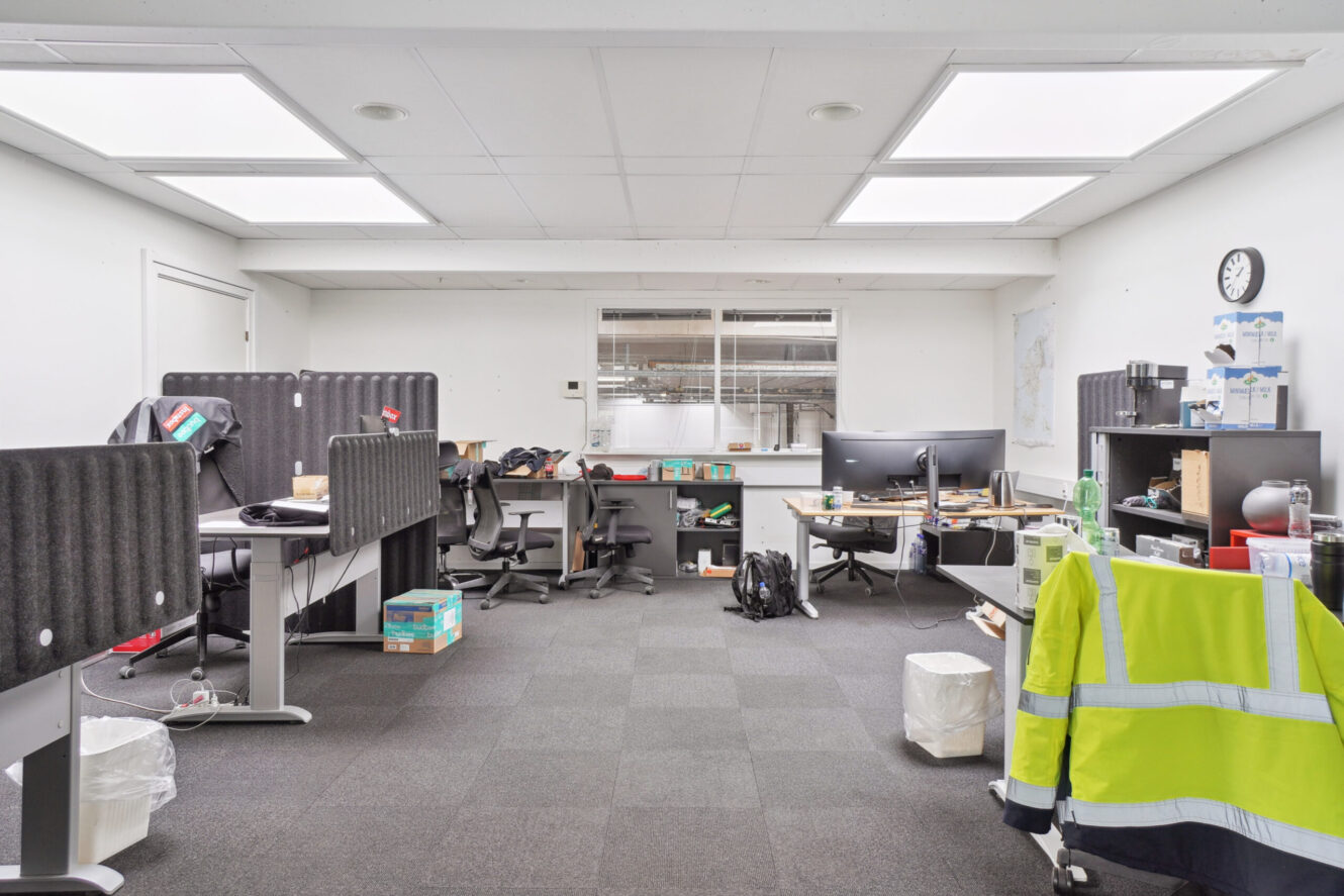 Et lyst kontor med seks skriveborde, computerskærme, kontorartikler, en sikkerhedsjakke på en stol og et vægur over hylderne til højre.