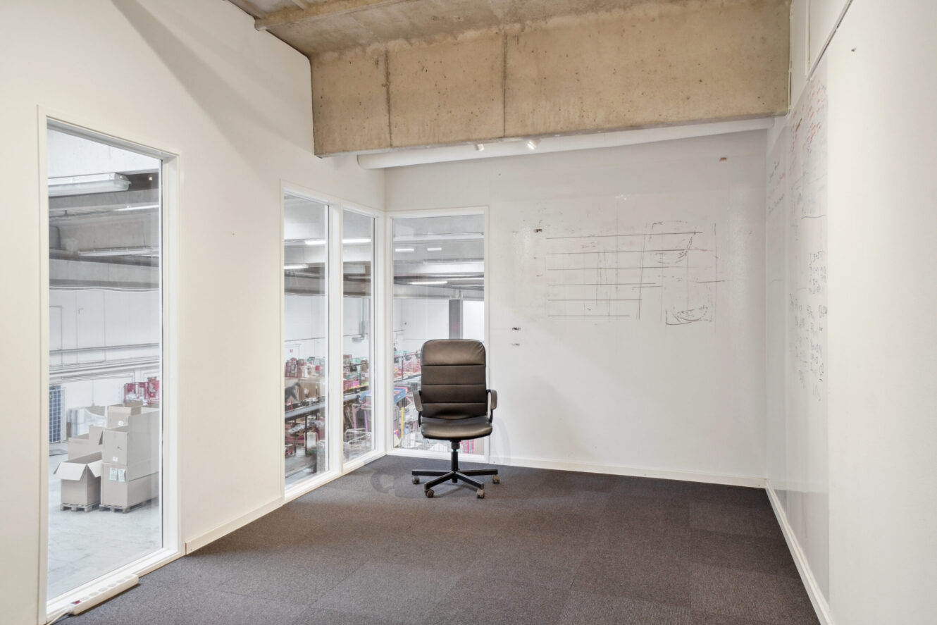 Et lille kontorlokale med en enkelt sort kontorstol, glasvinduer med udsigt til et lagerområde og whiteboards med svage markeringer på væggene.