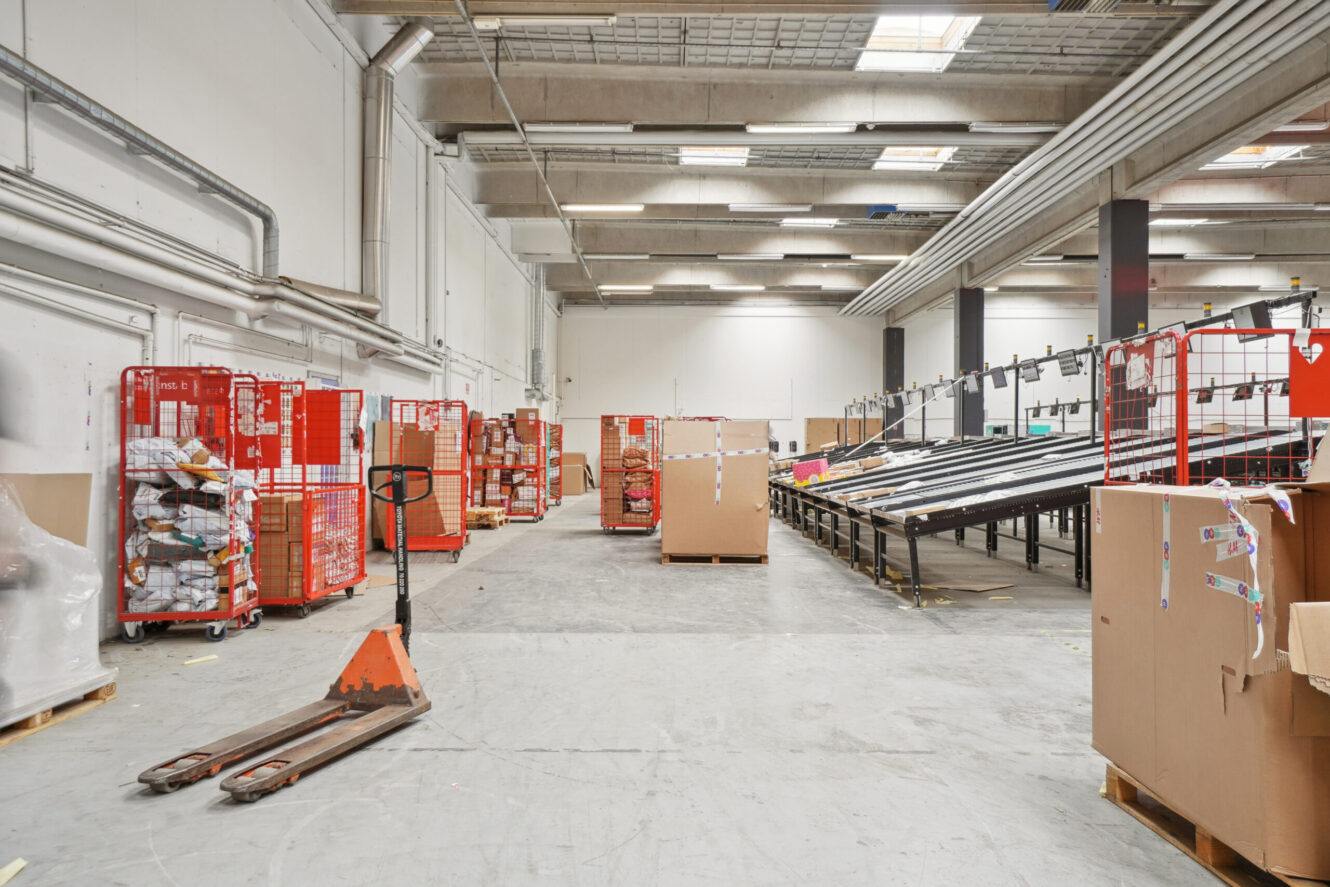 Et rummeligt lagerinteriør med røde metalvogne, papkasser, palleløftere og transportbånd under kraftig ovenlys.