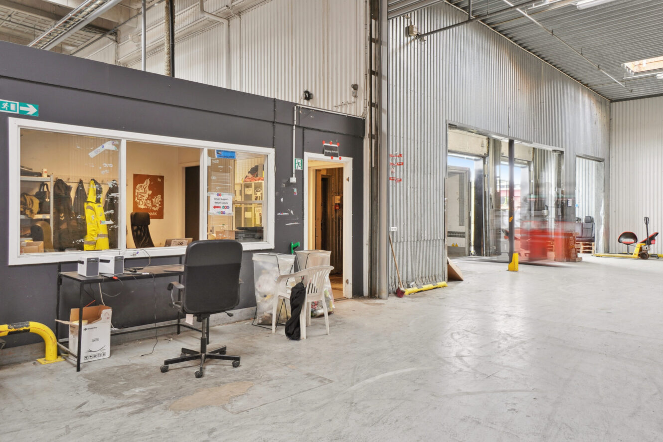 Et industrielt lagerinteriør med et kontorområde, skrivebord, stole og forskelligt udstyr; man kan se en gaffeltruck i bevægelse nær de åbne læsseramper.