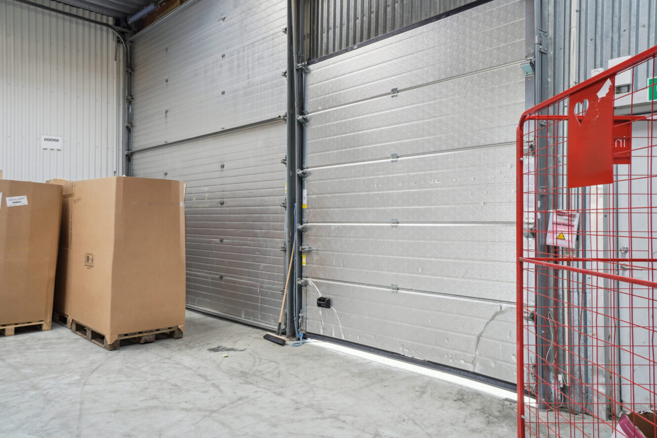 Et lagerområde med store lukkede metaldøre, to papkasser på paller, en kost og et rødt metalbur.