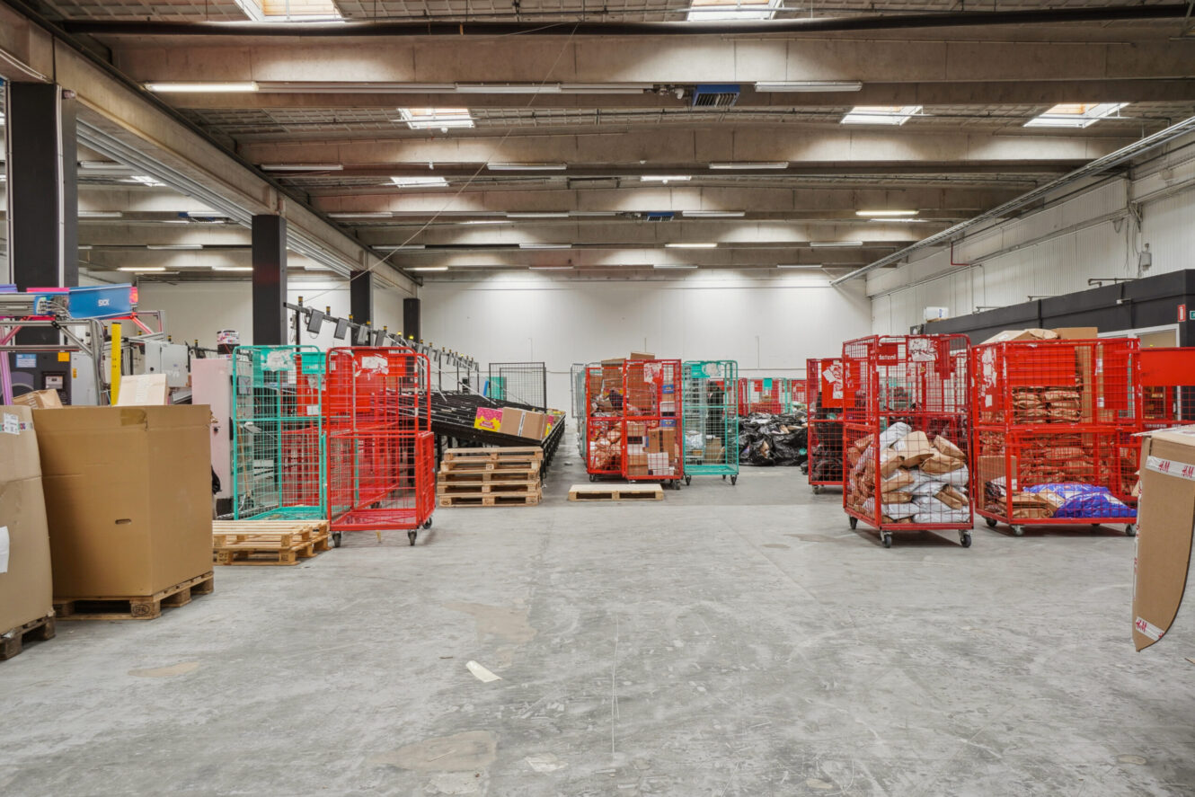 Rummeligt lagerinteriør med paller, papkasser og røde og grønne metalrullebure fyldt med forskellige varer under fluorescerende belysning.