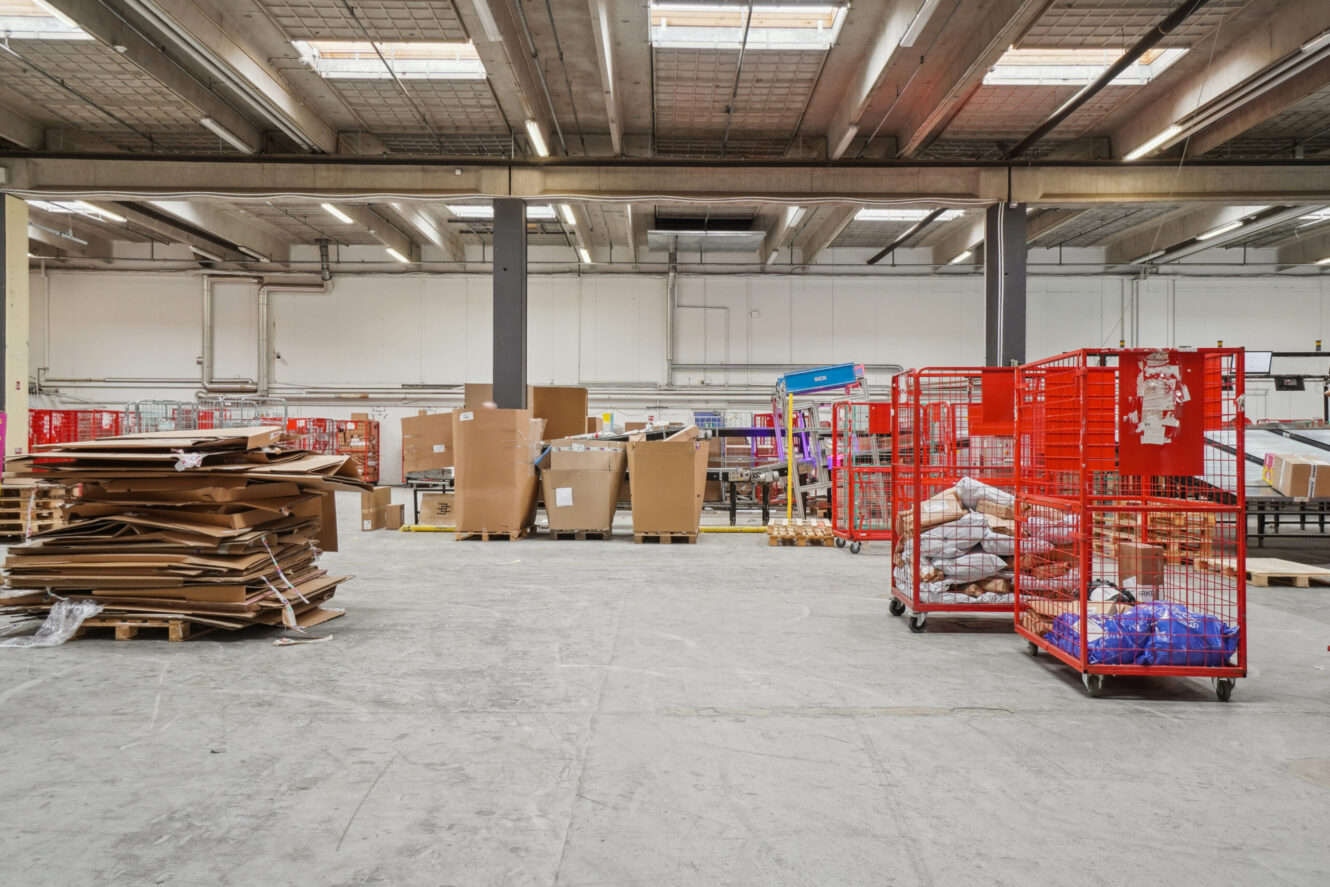 Et lagerinteriør med stabler af pap, paller, kasser og røde metalvogne fyldt med pakker på et betongulv under et højt loft.