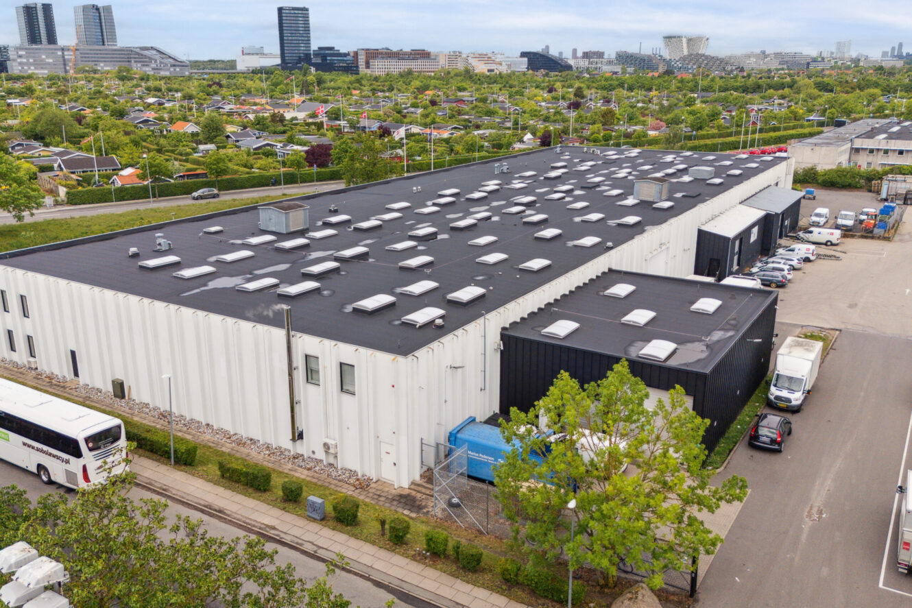 Stort industrilager med fladt sort tag og flere ovenlysvinduer, omgivet af grønne områder og nabobygninger; byens skyline ses i baggrunden.