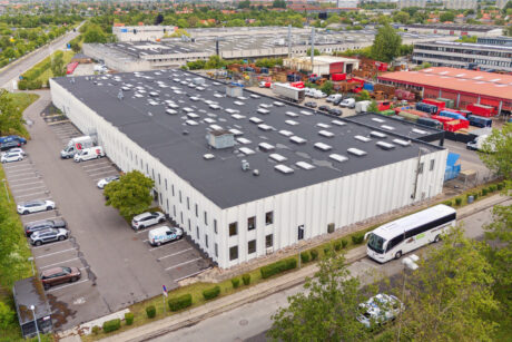 En stor hvid industribygning med fladt tag, omgivet af parkerede biler og en bus, beliggende i et byområde med andre erhvervsbygninger i nærheden.