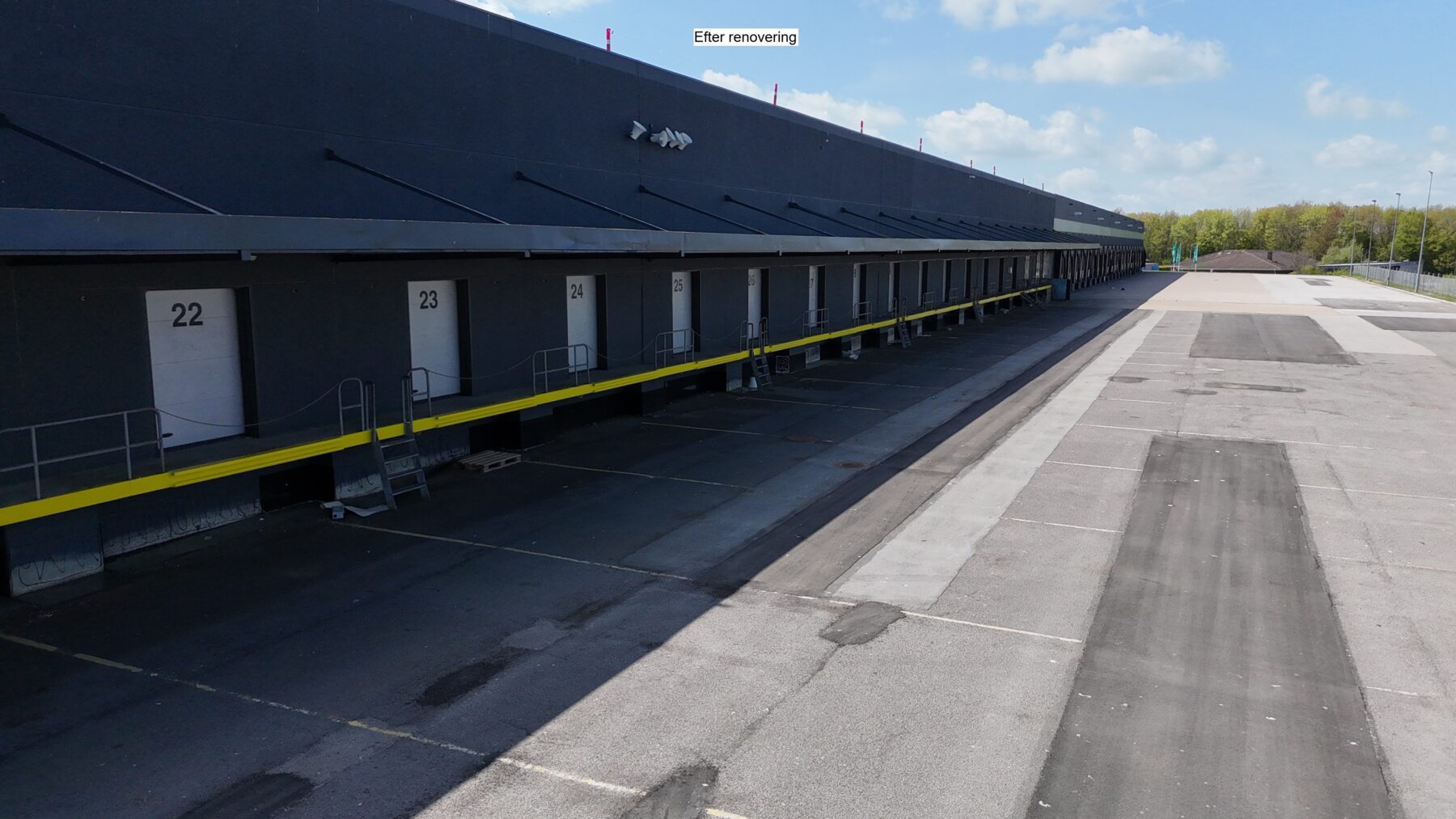 En række læsseramper med numrene 22 til 28 på en moderniseret lagerbygning med et tomt asfalteret område foran under en delvist overskyet himmel.