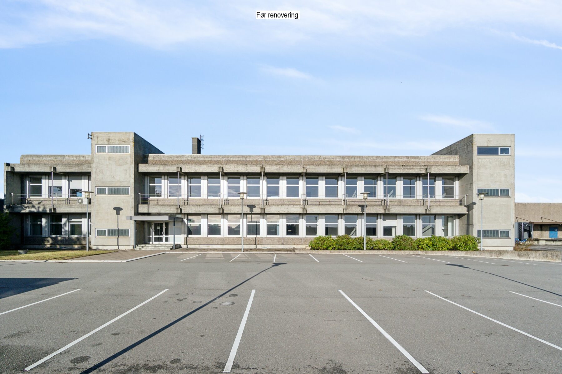 En toetagers betonbygning med mange vinduer, set fra en tom parkeringsplads under en klar himmel. På et skilt på taget står der "Til renovering".