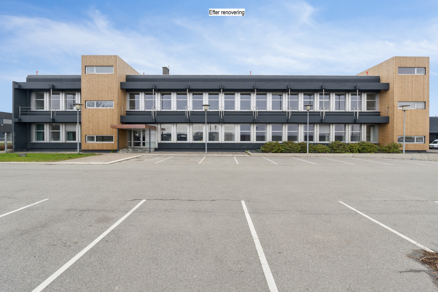 En toetagers kontorbygning med moderne facade, store vinduer og en tom parkeringsplads foran under en delvis overskyet himmel.