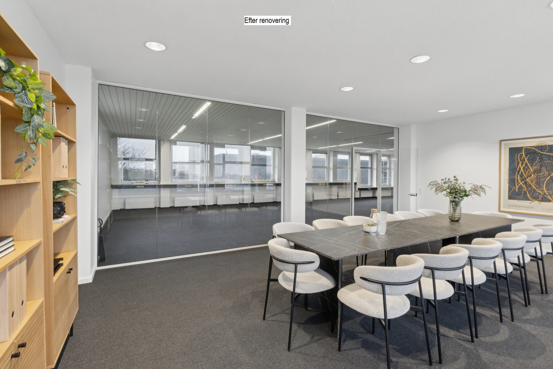 Moderne konferencerum med et langt sort bord, tolv hvide stole, glasvæg, hylder med planter og abstrakt kunst på væggen. På skiltet ovenover står der Efter renovering.