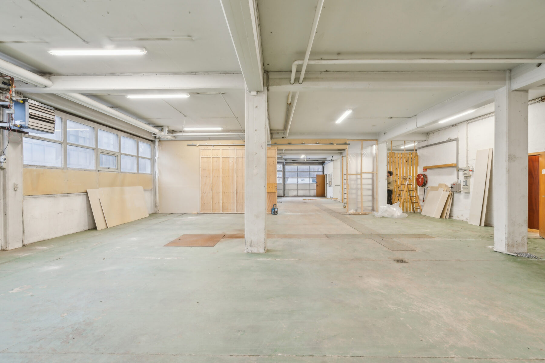 Rummelig, tom industrilagerbygning med betongulv, synlige søjler, store vinduer og lysstofrør. Nogle træpaneler og udstyr er placeret langs væggene.