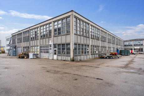 En stor, toetagers industribygning med mange vinduer og et ydre af beton, omgivet af en tom parkeringsplads under en delvis overskyet himmel.