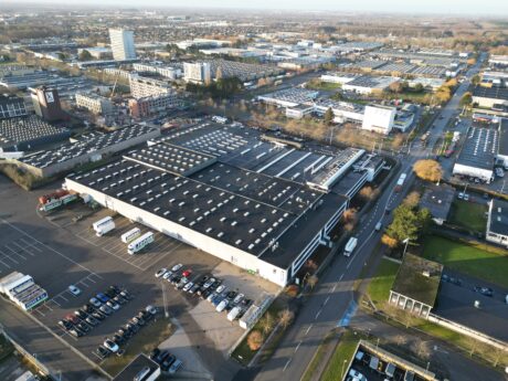 Luftfoto af et industriområde med store lagerbygninger, parkerede køretøjer og omkringliggende veje i et bymiljø.
