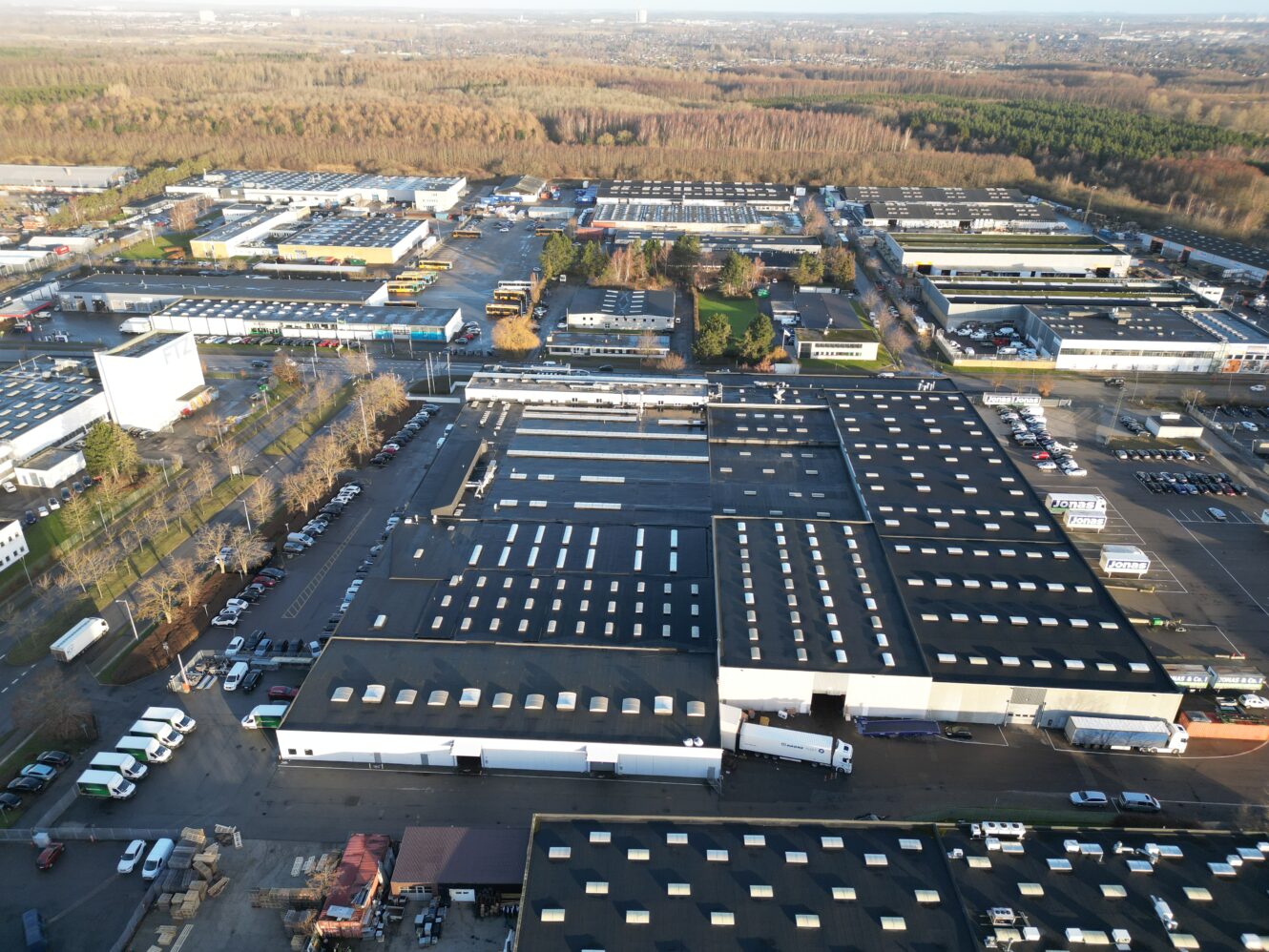 Luftfoto af et industriområde med store lagerbygninger, parkerede lastbiler, biler og omkringliggende veje, afgrænset af træer og skov i baggrunden.