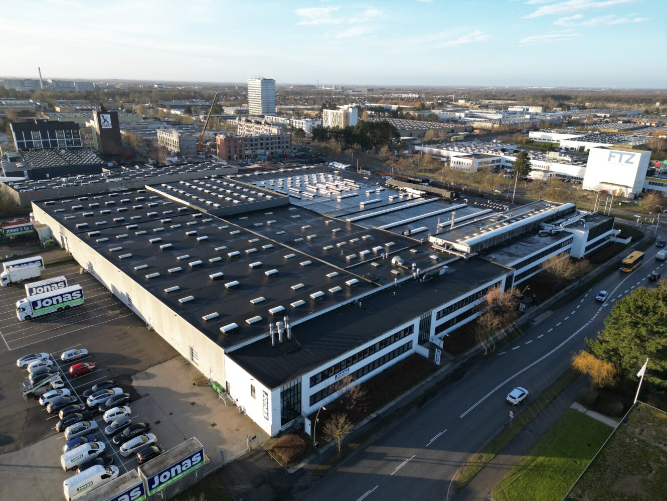 Luftfoto af en stor industribygning med parkeringsplads, omgivet af veje og bymæssig infrastruktur i baggrunden.