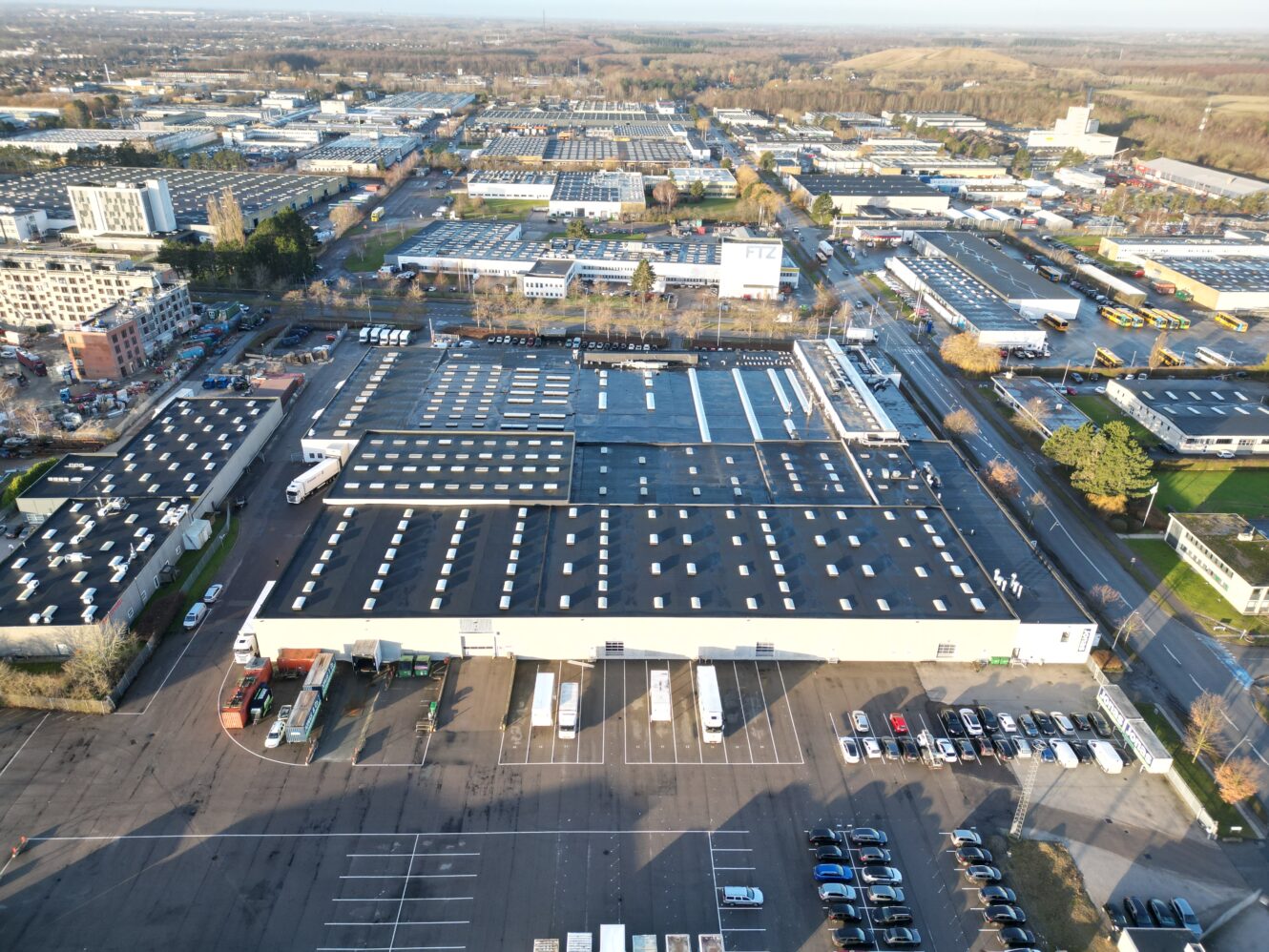 Luftfoto af et stort industrilager med lastbiler og biler parkeret udenfor, omgivet af flere bygninger og veje i et industriområde.