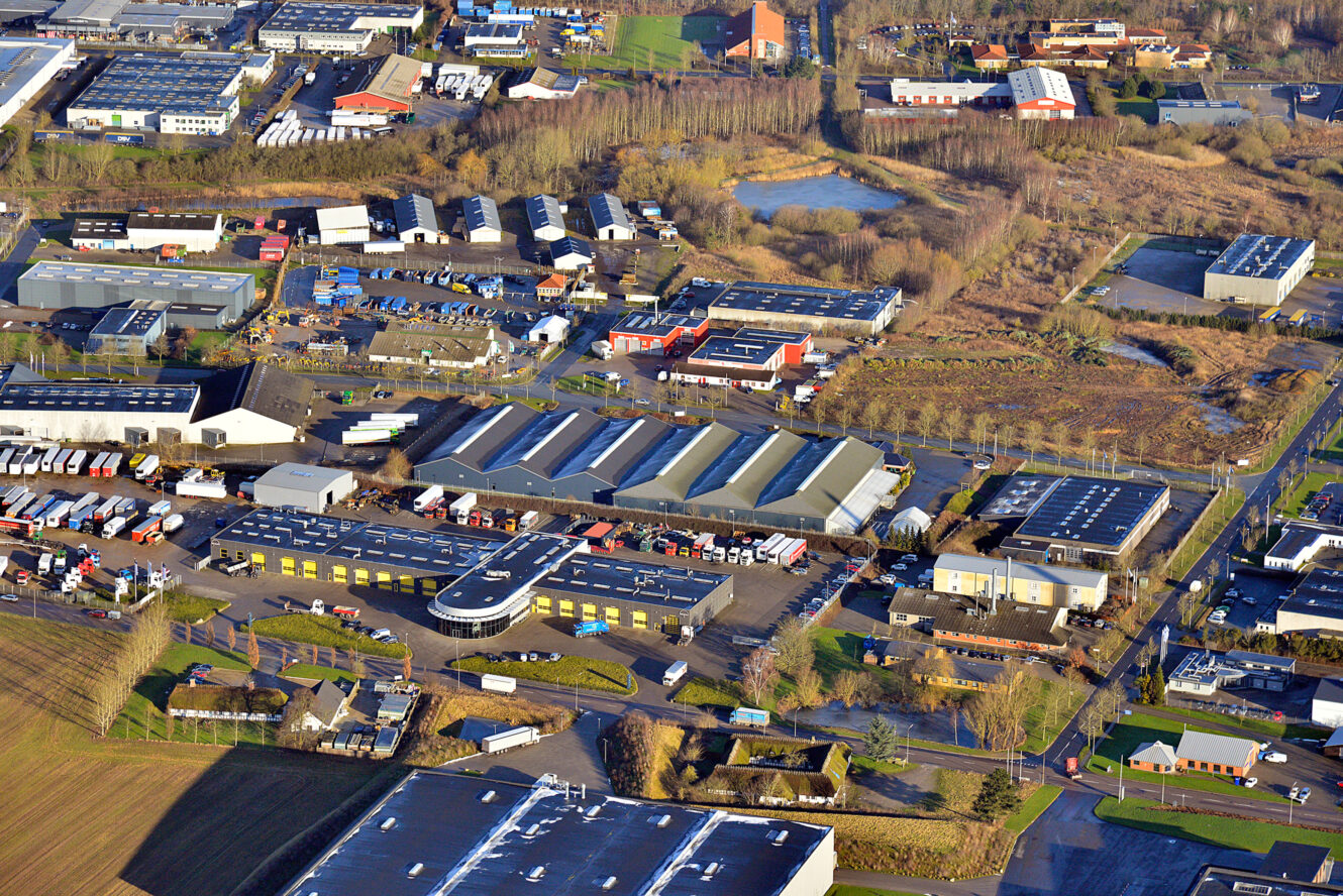 Luftfoto af et industriområde med lagerbygninger, erhvervsbygninger, parkerede lastbiler og pletter med græs og træer.