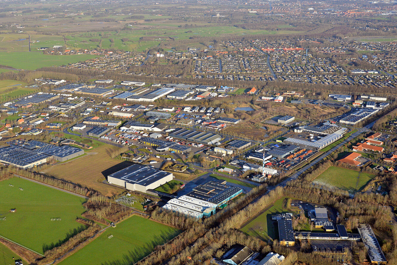Luftfoto af et industriområde med lagerbygninger, fabrikker og veje, omgivet af grønne marker og boligkvarterer i baggrunden.