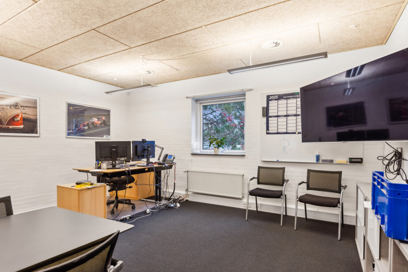 Lille kontor med skriveborde, computere og skærme, væghængt tv, kalender, stole, fotos på væggene og vindue med udsigt til det fri.