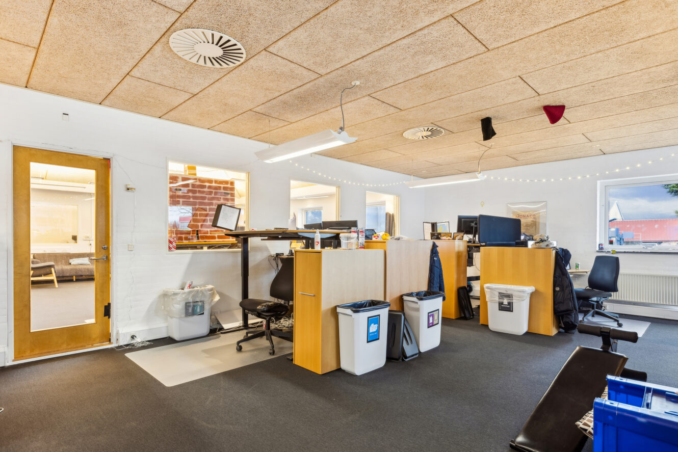 Moderne kontorlokaler med træborde, computere, genbrugsspande og et hæve-sænkebord; lyse vægge og lofter, lyskæder og vinduer, der giver naturligt lys.