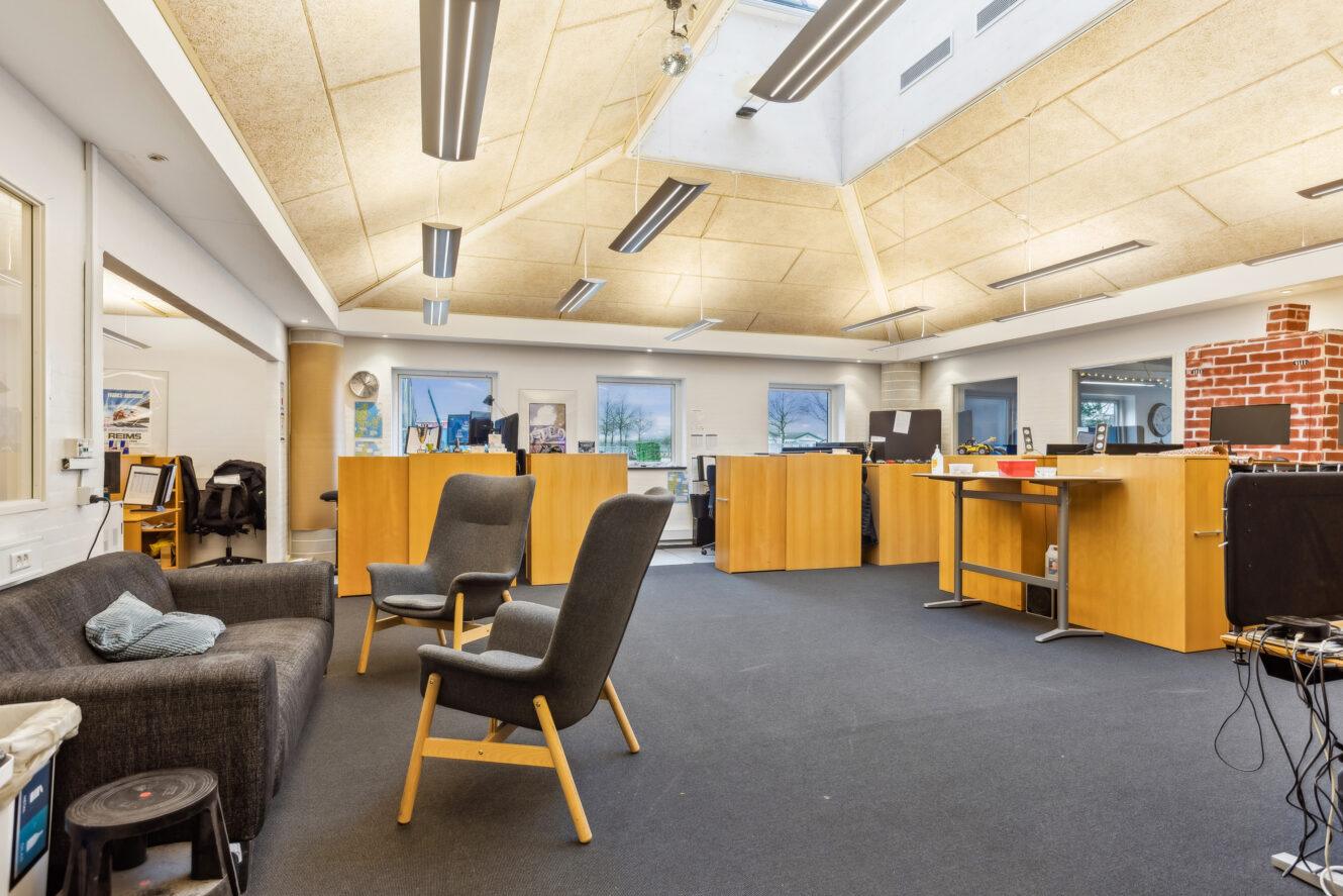 Moderne kontorlokaler med ovenlys, sort gulvtæppe, træborde, to lænestole, en sofa og computere på skriveborde.