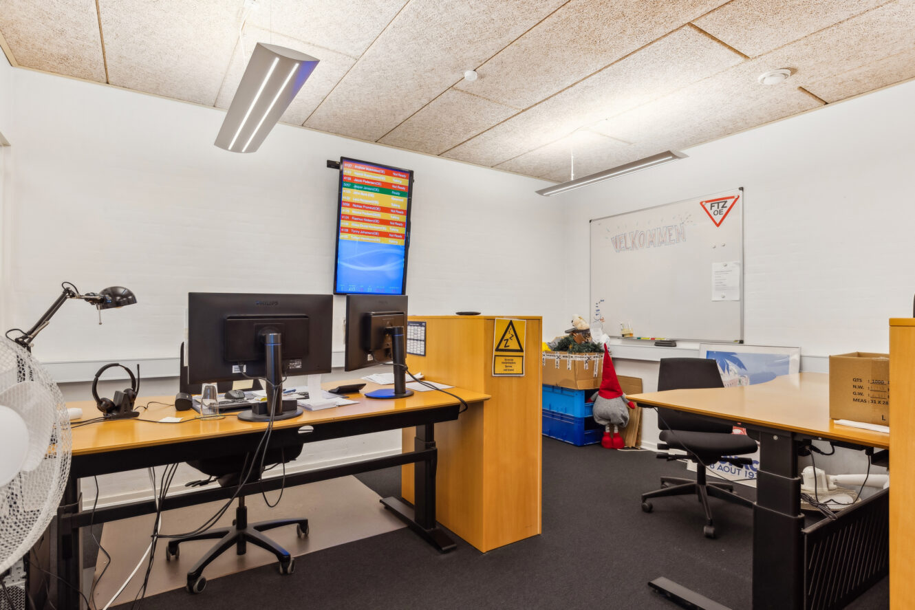 Moderne kontor med to skriveborde, computerskærme, en væghængt skærm, der viser data, et whiteboard med VELKOMMEN skrevet på og diverse kontorartikler og udstyr.