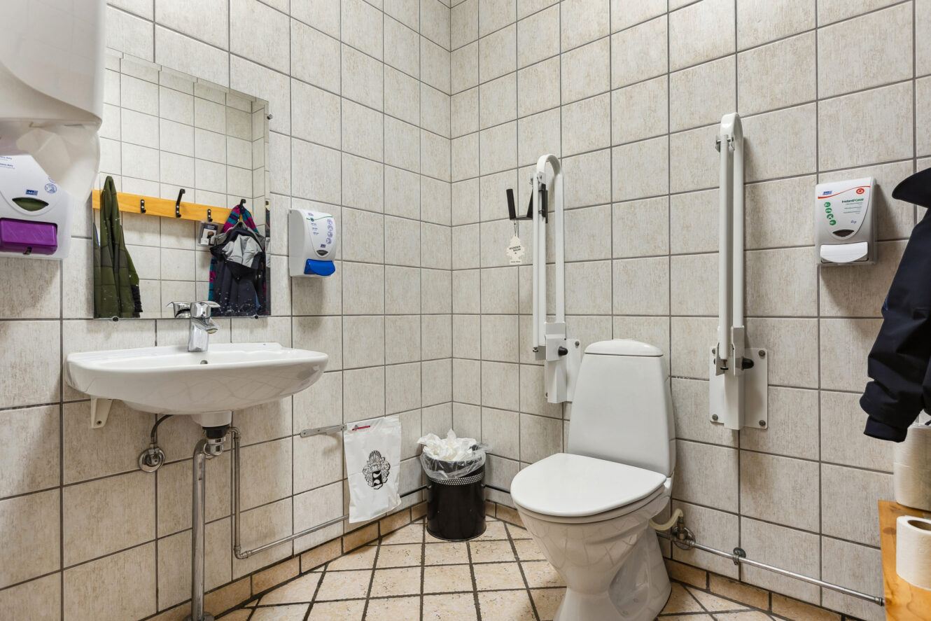 Tilgængeligt offentligt toilet med hvide flisevægge, toilet med støtteben, håndvask, spejl, sæbedispensere, knager, affaldsspande og synlige toiletartikler.