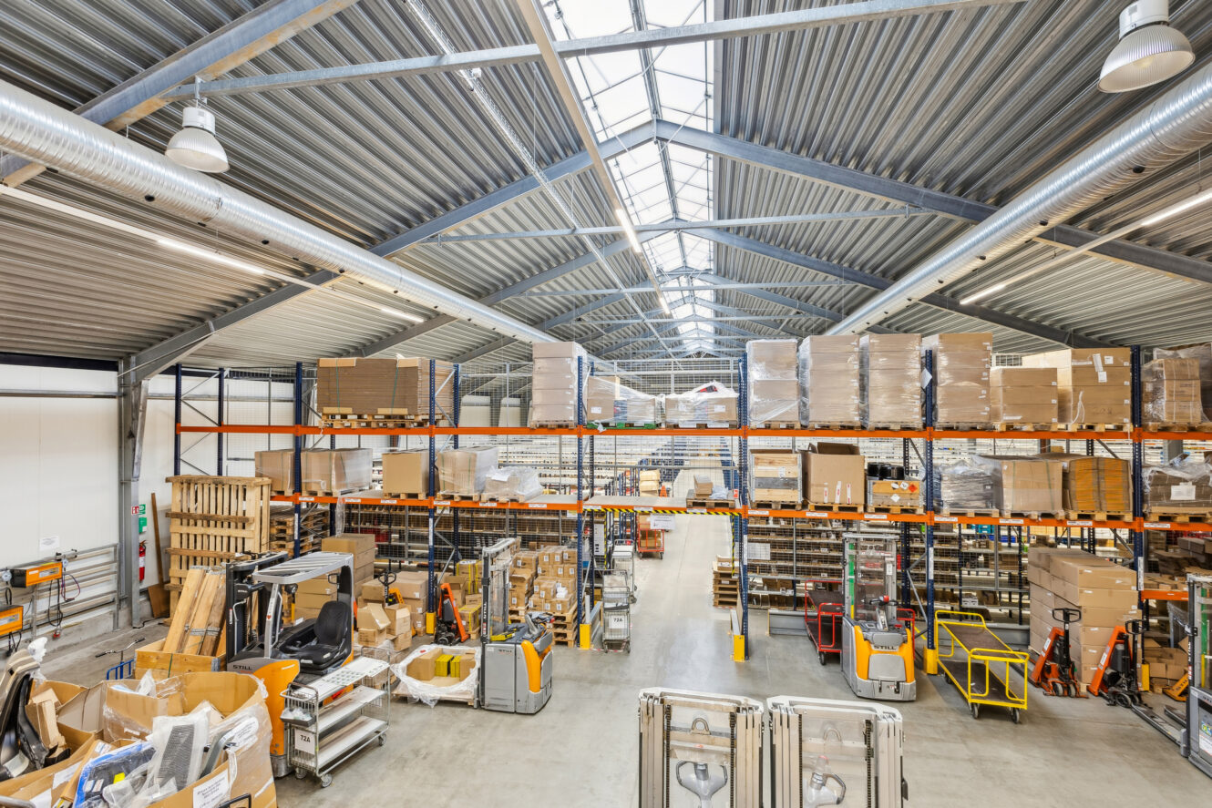 Stort lagerinteriør med hylder fyldt med kasser og paller, forskelligt udstyr og vogne spredt på betongulvet og ovenlysvinduer, der lukker naturligt lys ind.