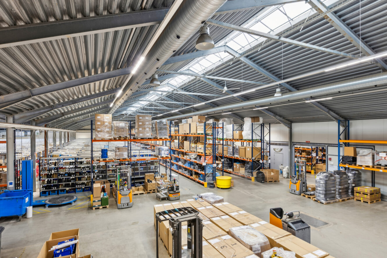Stort lagerinteriør med høje metallofter, hylder fyldt med kasser og produkter, paller på gulvet og kraftig industriel belysning.