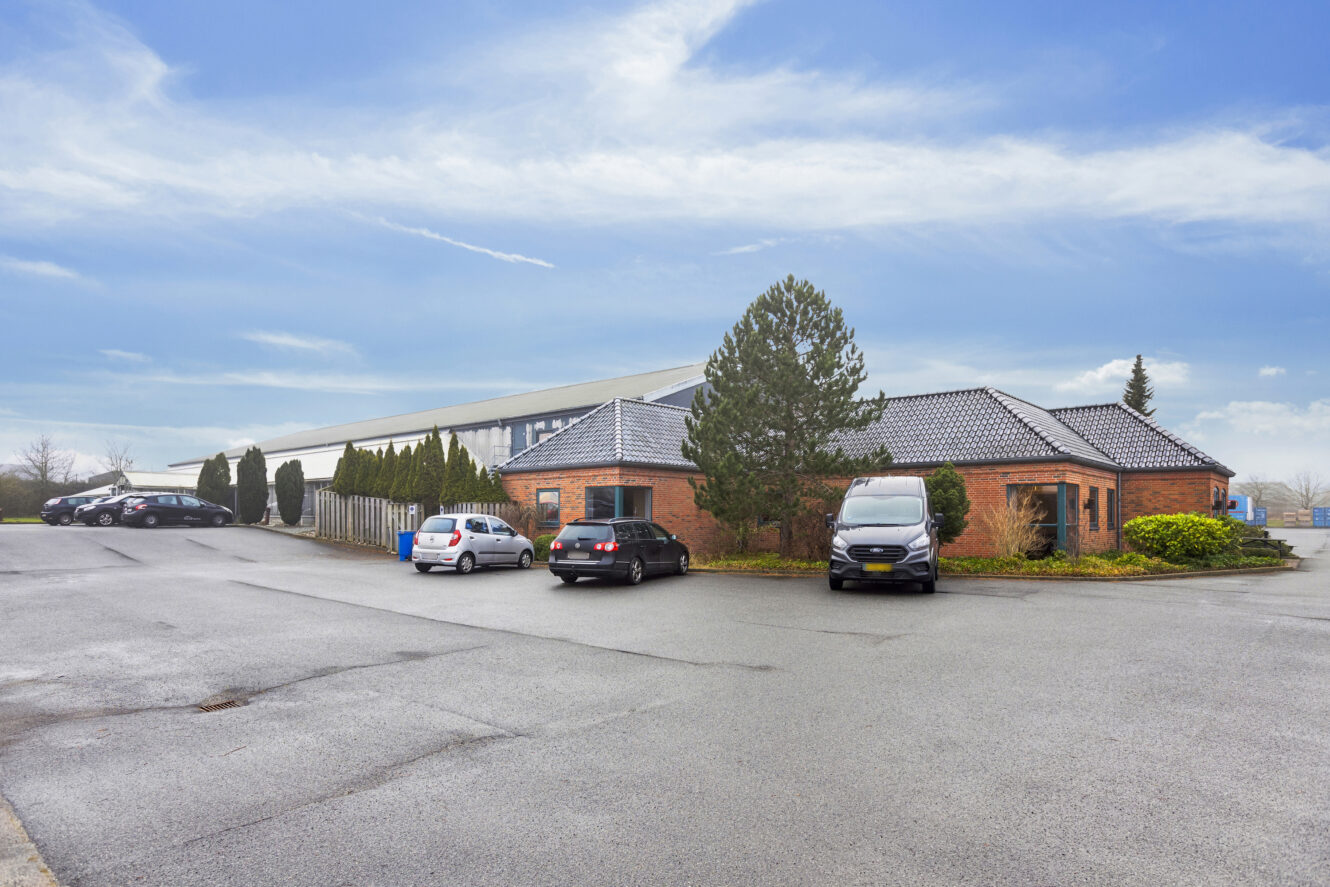 En kontorbygning i mursten med flere parkerede biler foran, beliggende på en stor asfalteret grund under en delvis overskyet himmel.