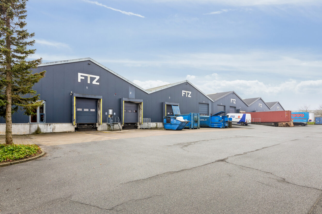 En række grå industrilagerbygninger med læsseramper mærket FTZ, containere og en skibscontainer udenfor under en overskyet himmel.