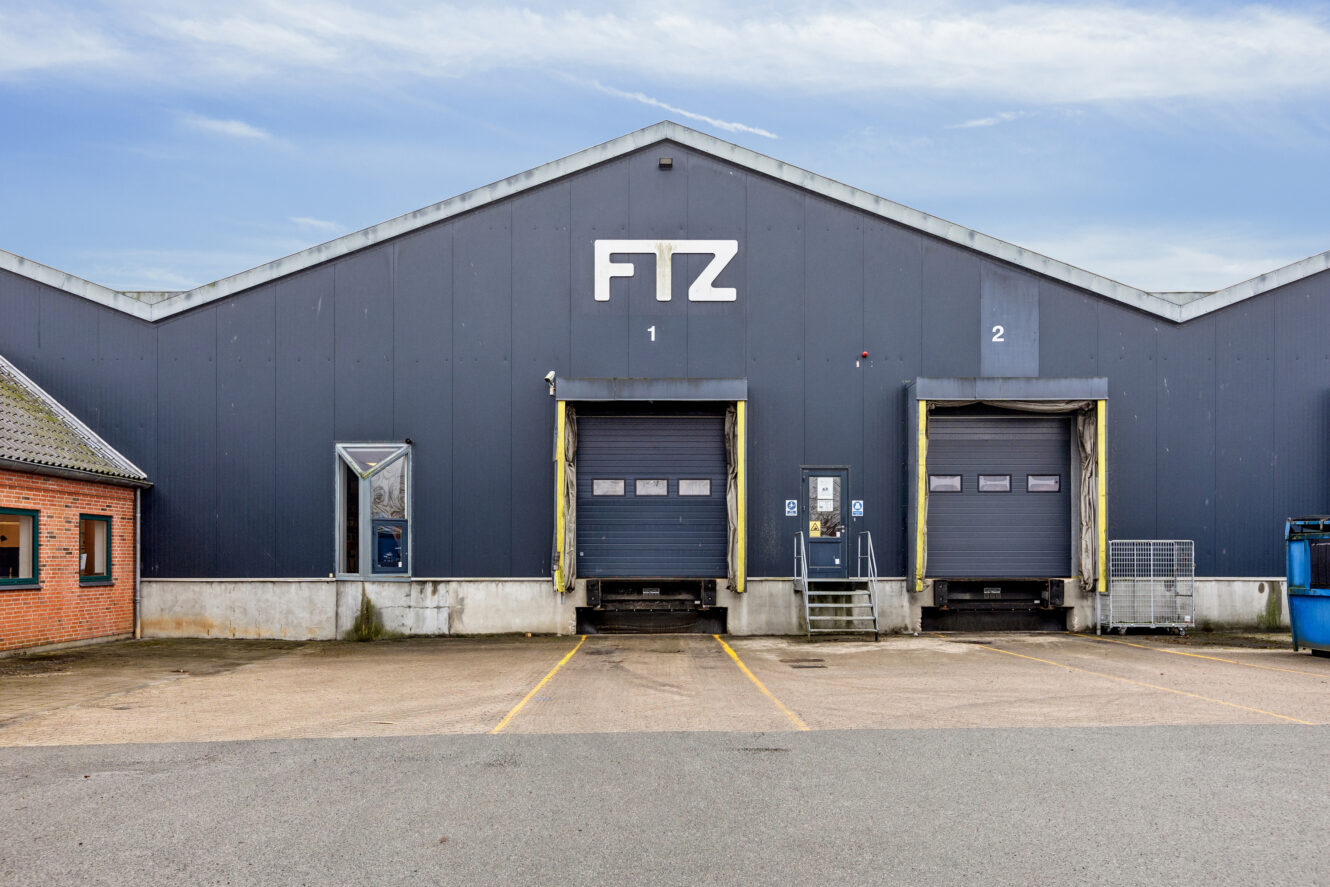Et gråt industrilager med FTZ på facaden, med to læsseramper nummereret 1 og 2, og en muret kontorsektion til venstre.
