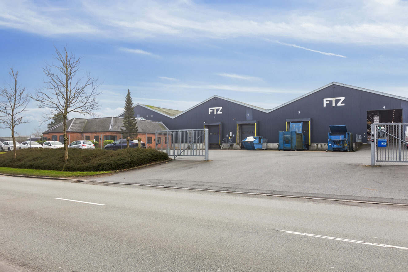 En stor industribygning med "FTZ"-skilte, indhegnet indgang, parkerede biler og blå affaldscontainere, set fra den anden side af gaden på en klar dag.