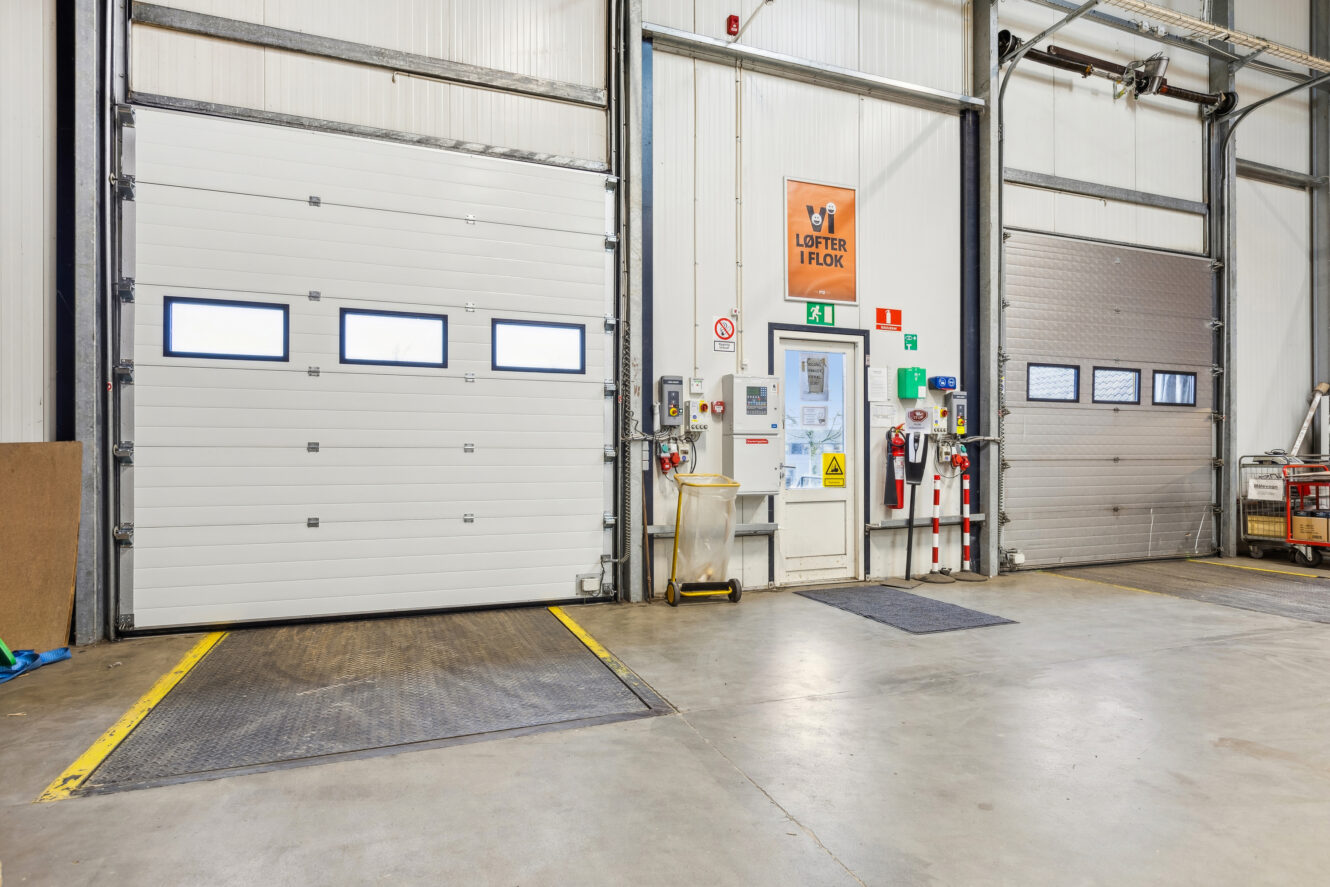 Industrielt læsserampeområde med to lukkede overliggende døre, sikkerhedsskiltning, udstyr og en personaleindgang mellem dørene.