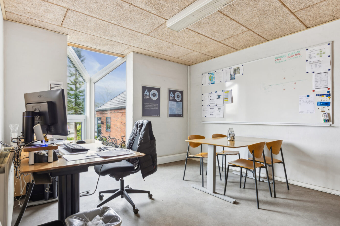 Moderne kontor med skrivebord, computer, drejestol, lille mødebord med fire stole, stort vindue og whiteboard med noter og diagrammer på væggen.